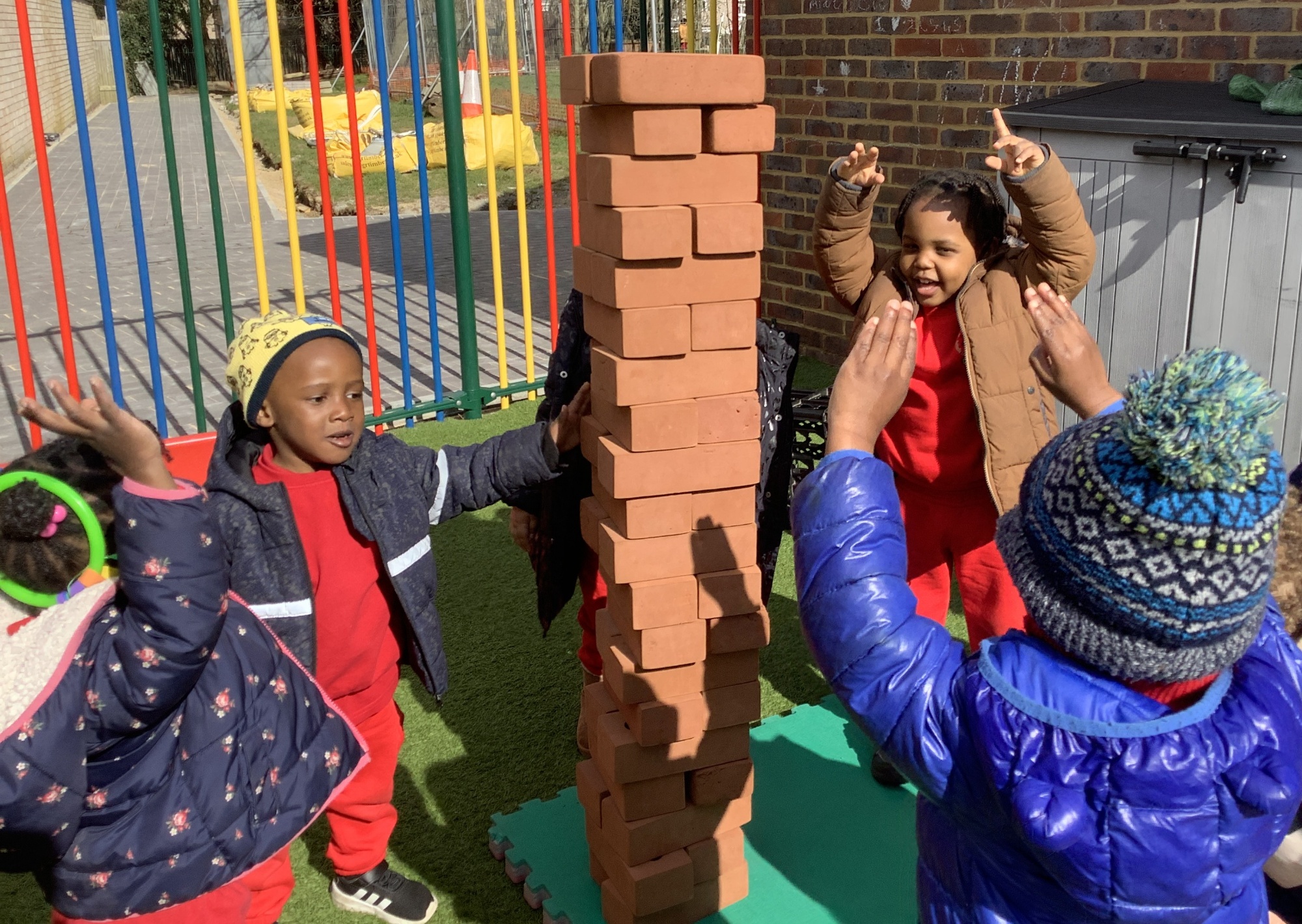 Young children are smiling at a tower they have built together from blocks in the outside nursery garden.